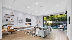 A modern living room with a grey sofa, wooden flooring, and a large window overlooking a garden. - LA Villa by Avantstay Walk to The Grove LA (Los Angeles)