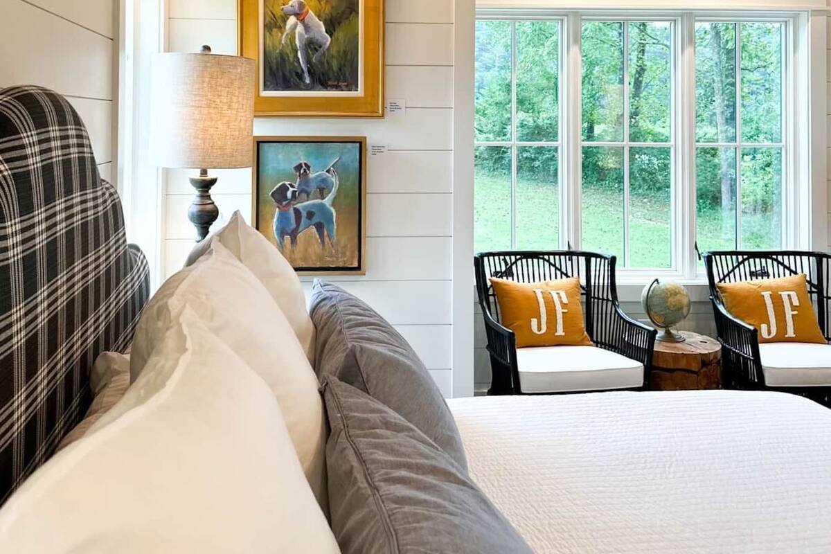 A bedroom with a bed, two chairs with 'JF' pillows, a mounted deer head, and two framed paintings.