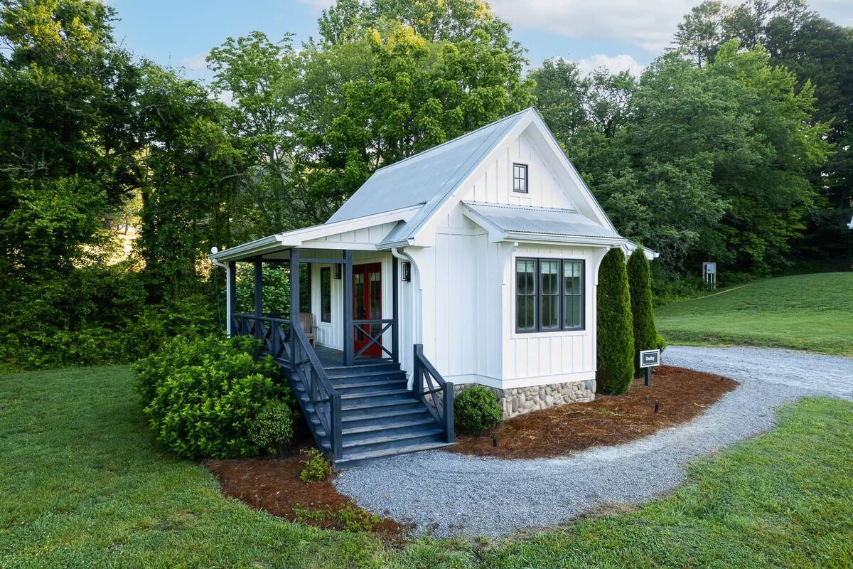 A small white cottage with a porch and steps, surrounded by greenery.