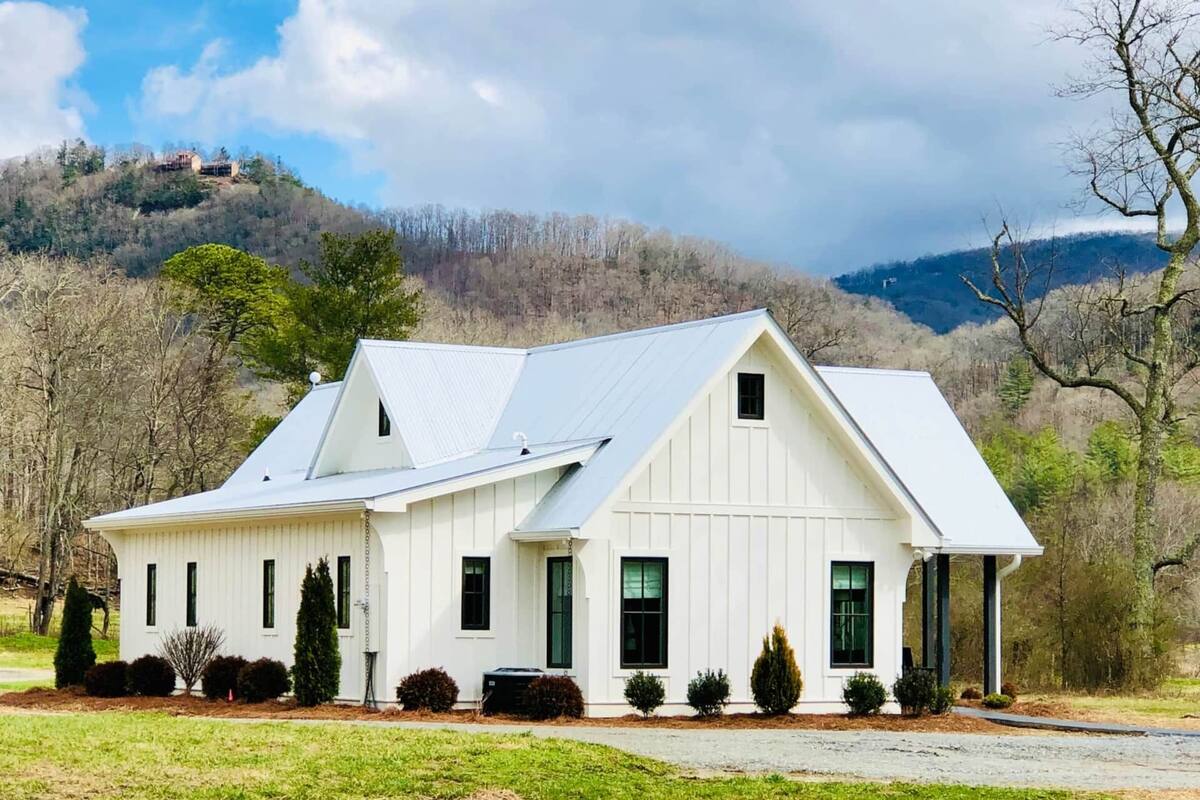 A white two-story house with a gabled roof, surrounded by a well-maintained lawn and a hilly landscape in the background.