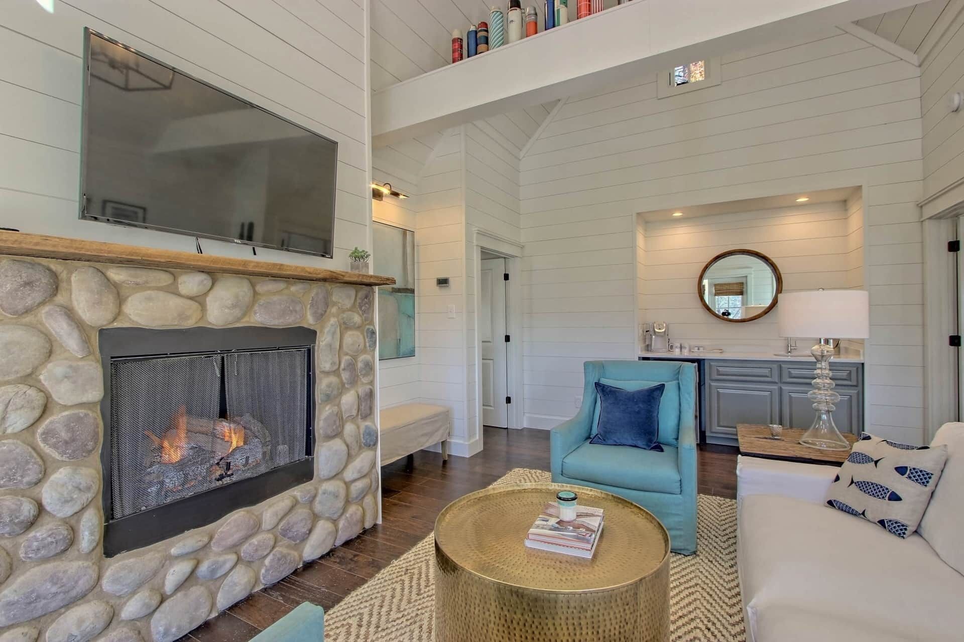A spacious living room with a stone fireplace, a large flat-screen TV mounted above it, and a cozy seating area with a blue armchair and a white sofa.