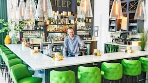 Un espace bar avec un barman, des tabourets de bar verts et une étagère bien garnie de différentes bouteilles et de verres.