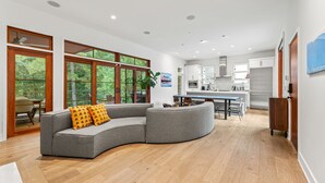 A modern interior with a curved grey sofa, wooden flooring, and a kitchen area with white cabinets and a stainless steel refrigerator. - The Red House in Serenbe w/ Golf Cart (Palmetto)