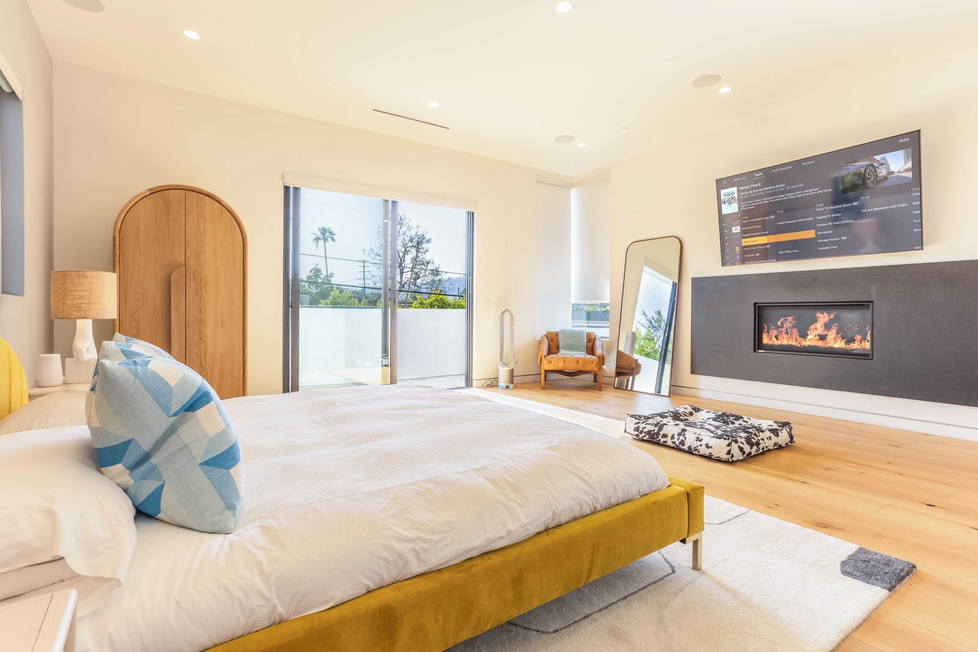 Een moderne slaapkamer met een groot bed, een open haard, een televisie en een schuifpui naar buiten.