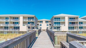 A wooden boardwalk leading to a row of multi-story buildings with balconies, likely a hotel or resort. - Starboard Village by Southern Vacation Rentals (Pensacola Beach)