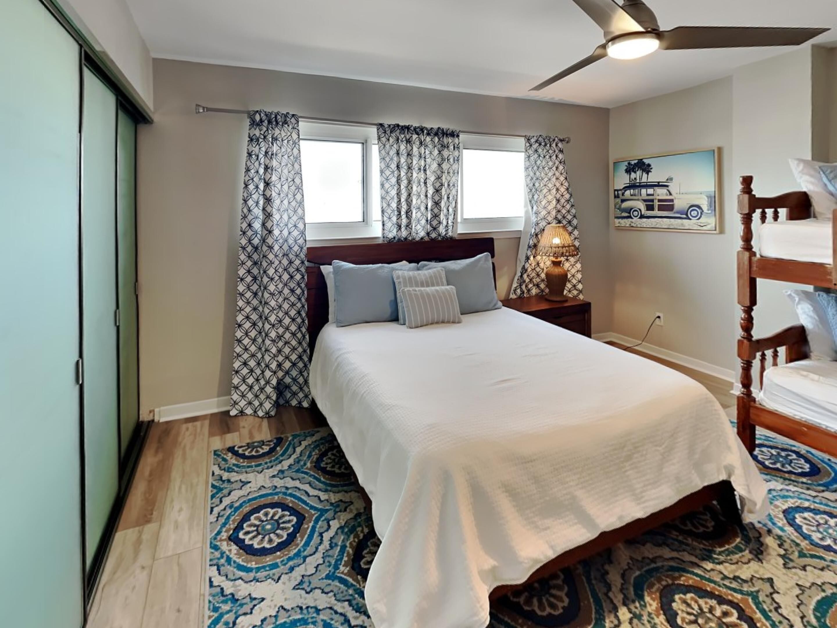 A bedroom with a bed, a nightstand, a lamp, a ceiling fan, a window with curtains, and a painting on the wall.