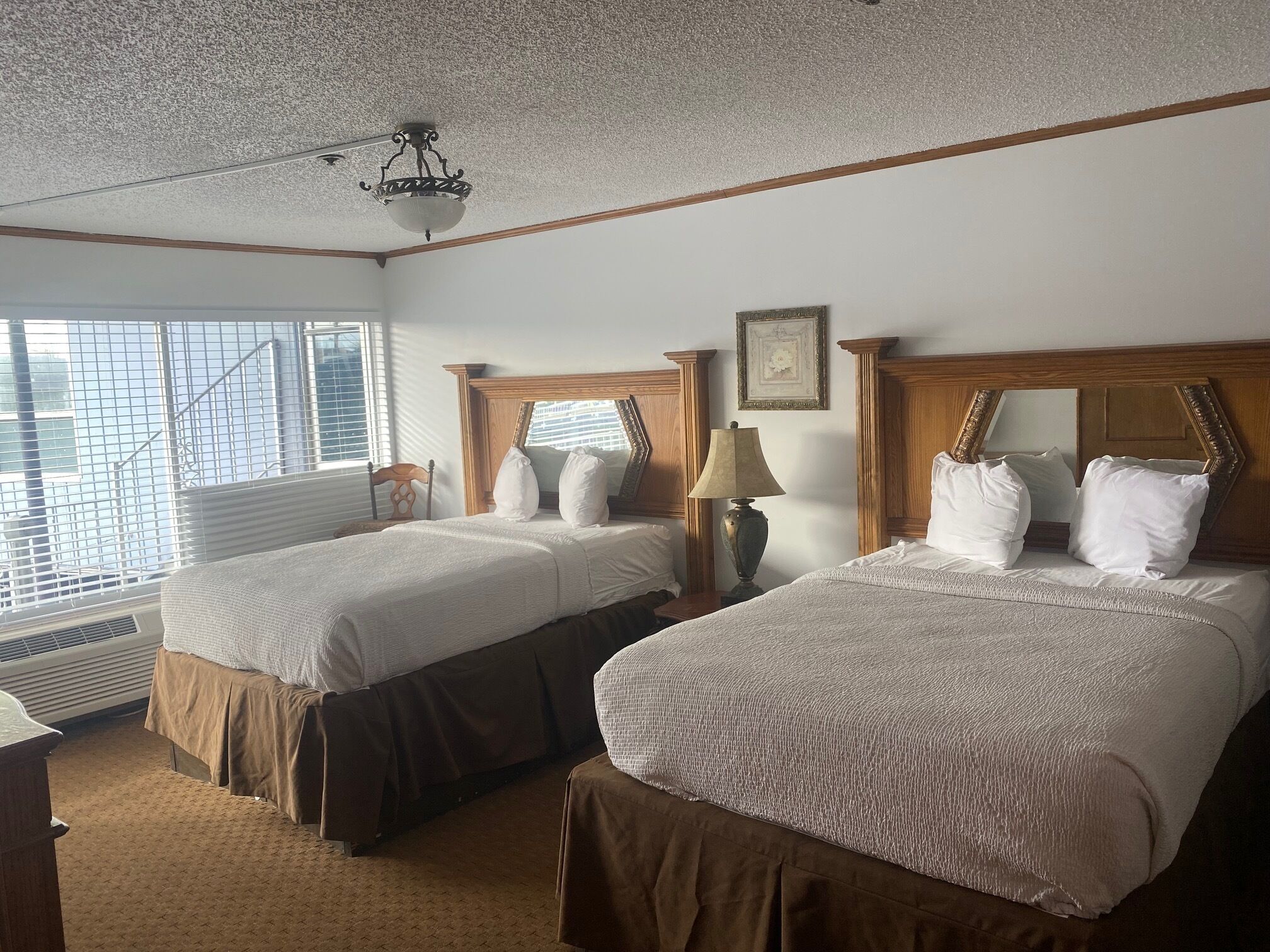 A hotel room with two beds, wooden headboards, a ceiling fan, a window with blinds, and a framed picture on the wall.