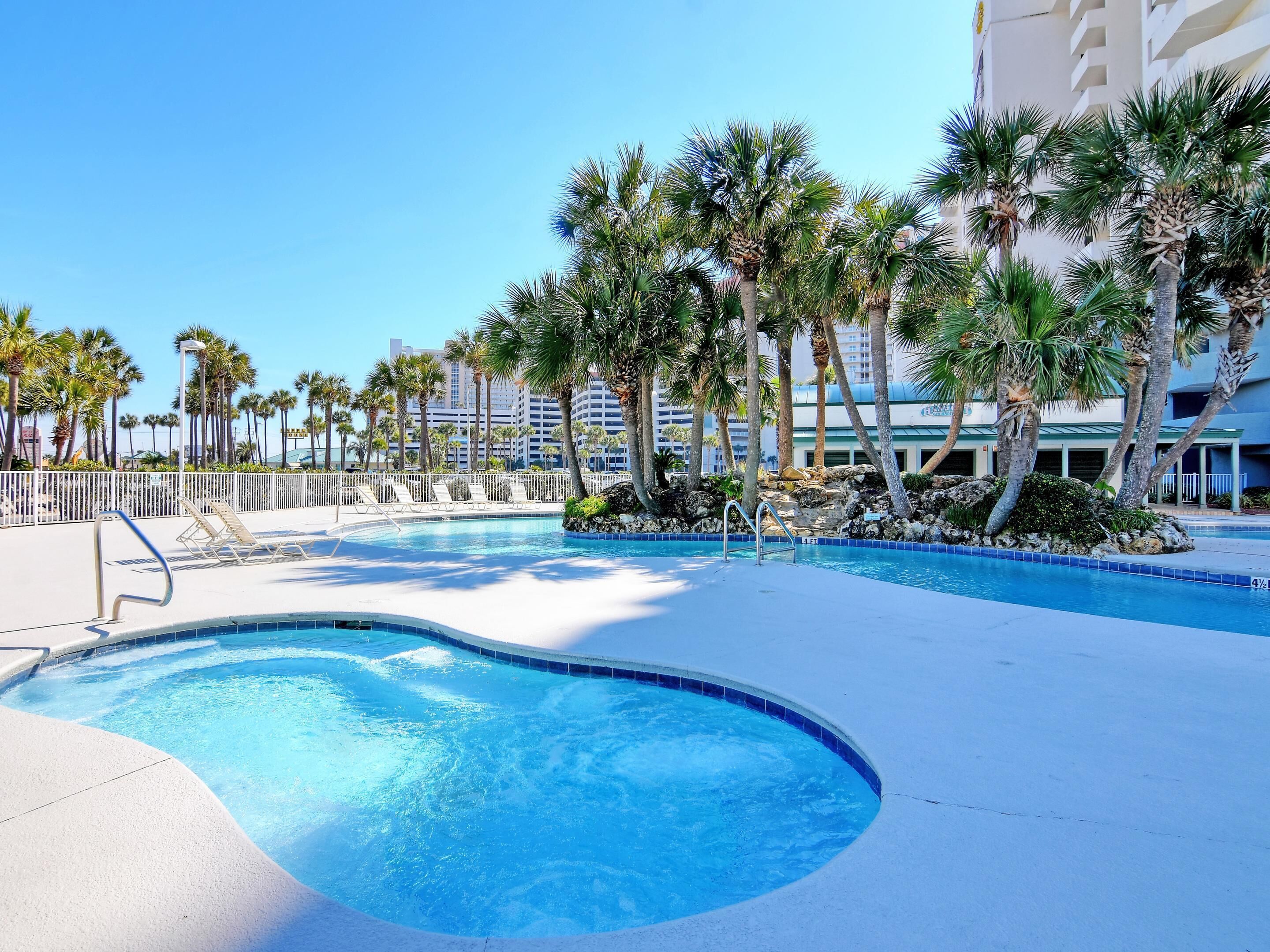 A swimming pool with lounge chairs and palm trees, surrounded by a white fence and buildings in the background.