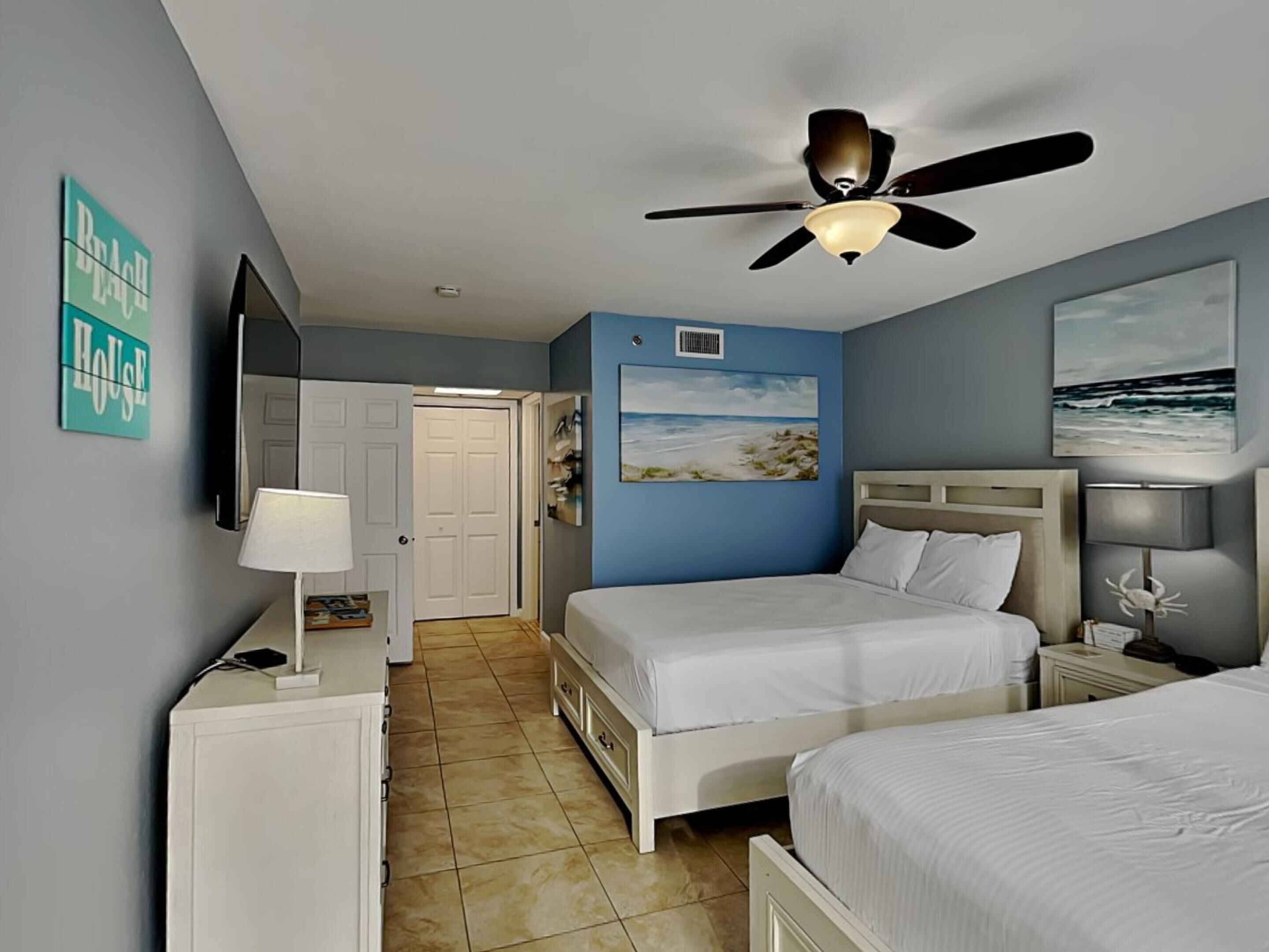 Habitación de hotel con dos camas, ventilador de techo, un cuadro de playa y una puerta que da a un baño.