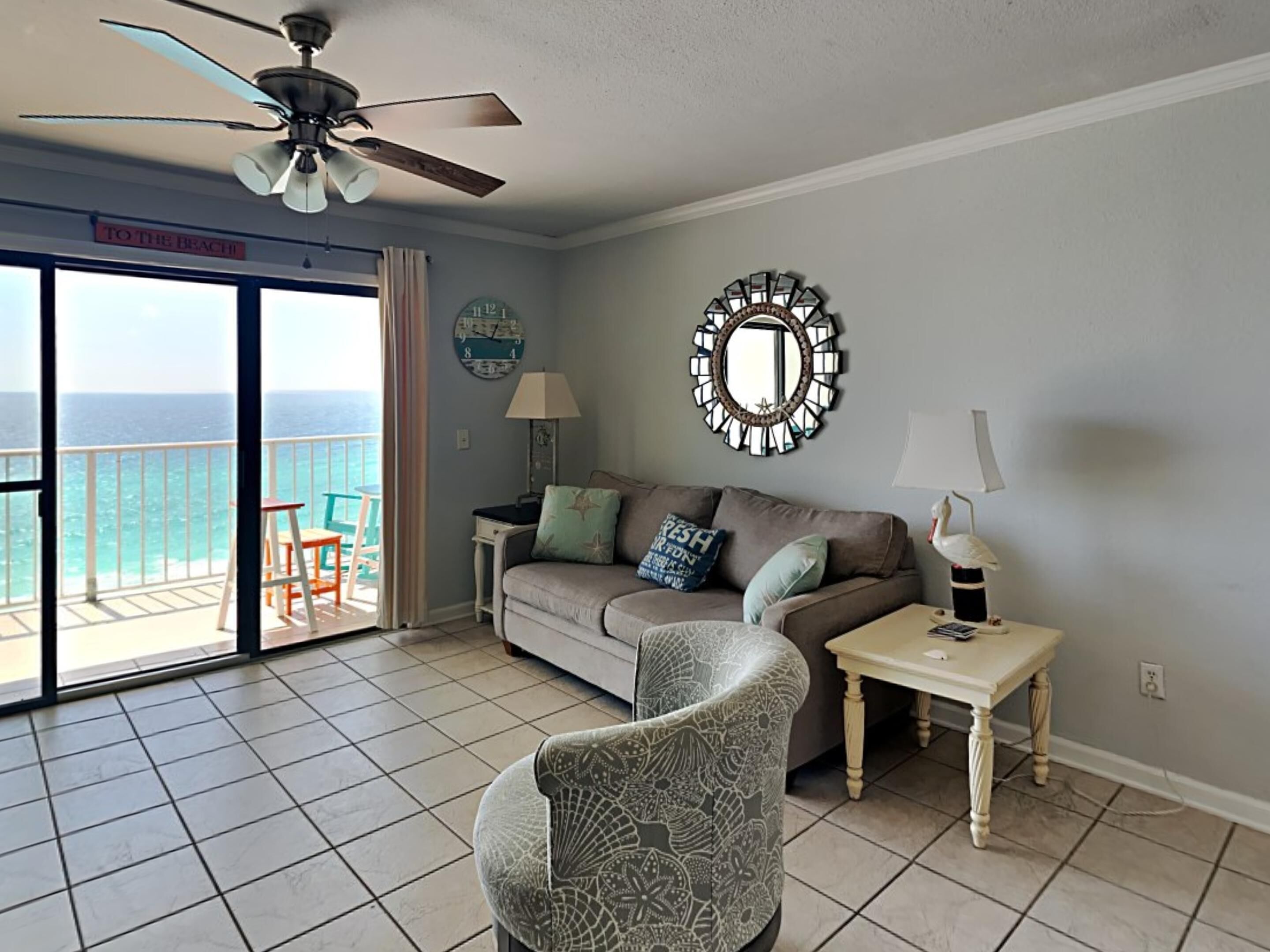 A living room with a balcony, a ceiling fan, a clock, a mirror, a lamp, and a table.