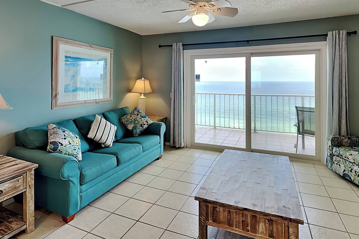 A living room with a teal sofa, a wooden coffee table, a ceiling fan, a framed picture, a lamp, and a view of the ocean through a sliding glass door.