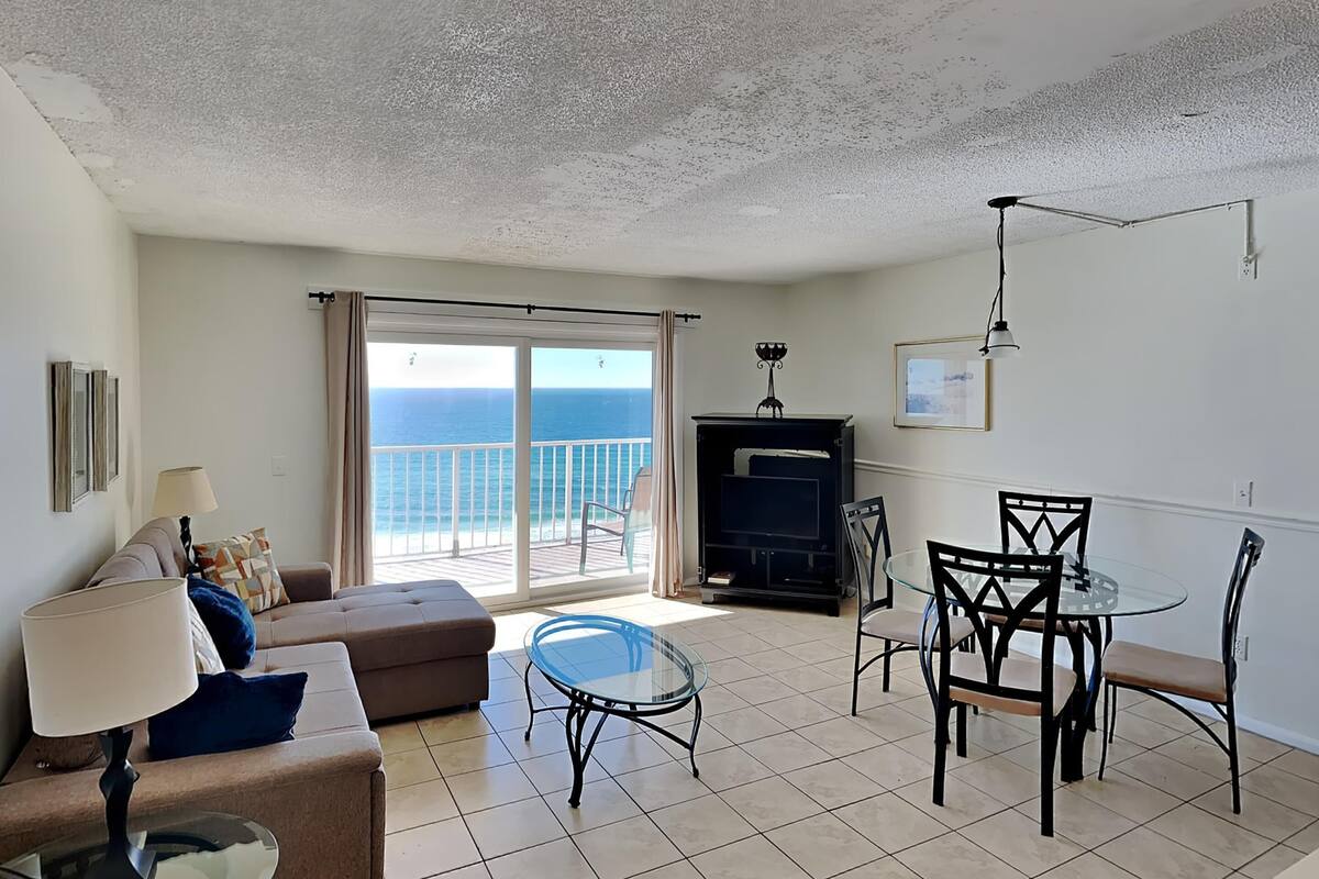A living room with a sofa, a glass table, and a dining area with chairs. There is a view of the ocean through a sliding door.