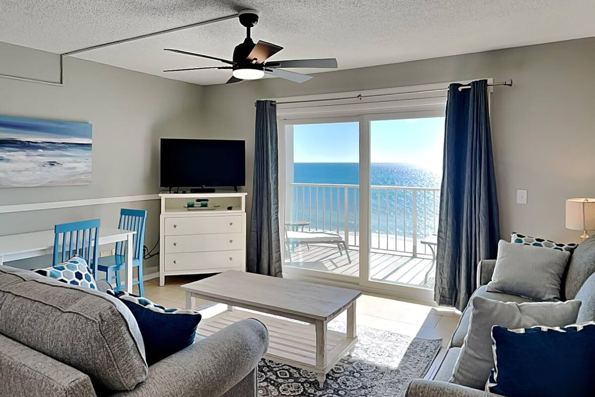A living room with a TV, a ceiling fan, a sofa, a coffee table, and a view of the ocean through large windows.