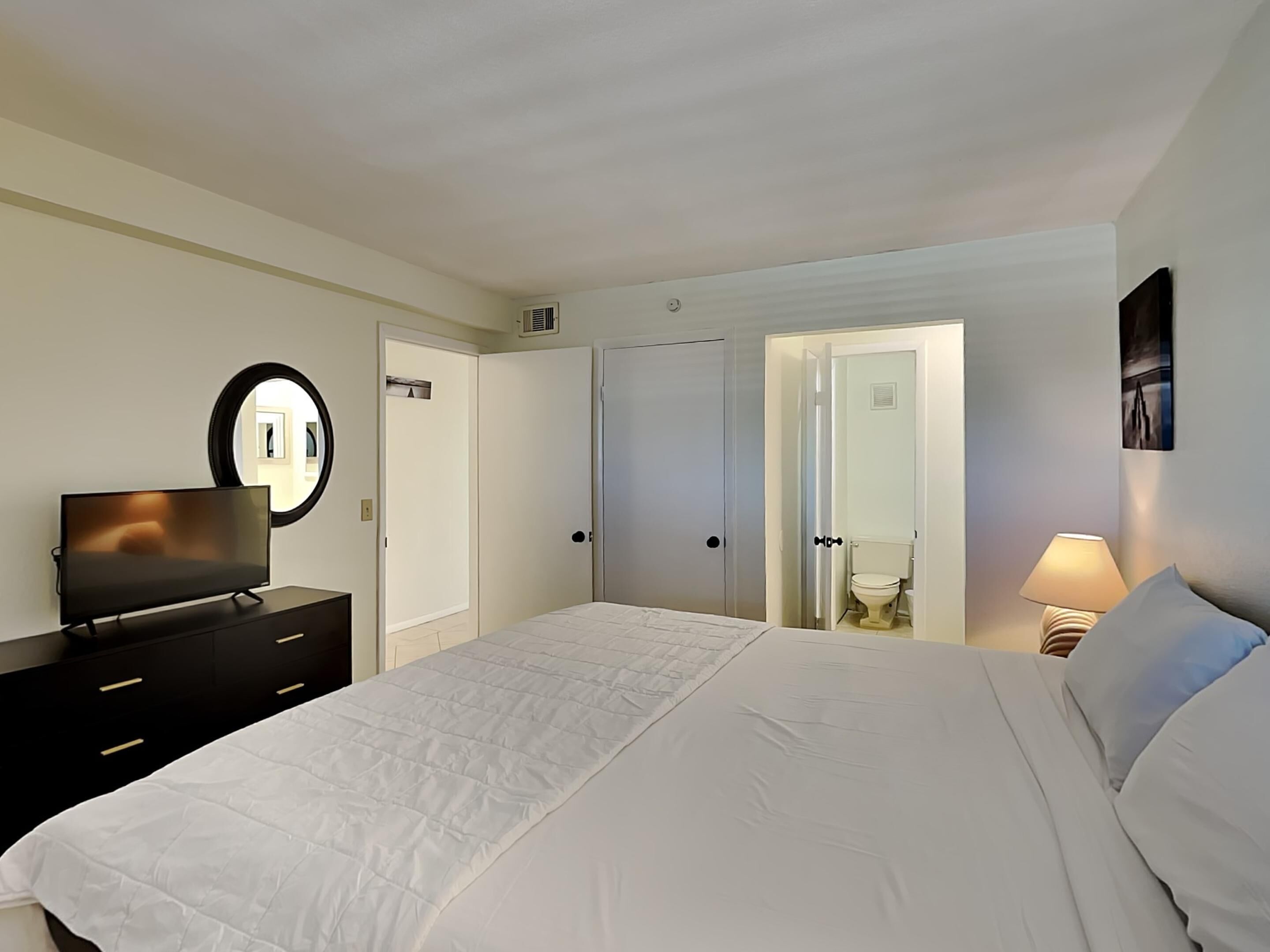 Un dormitorio con cama, televisión sobre una cómoda, espejo y puerta que da a un baño.