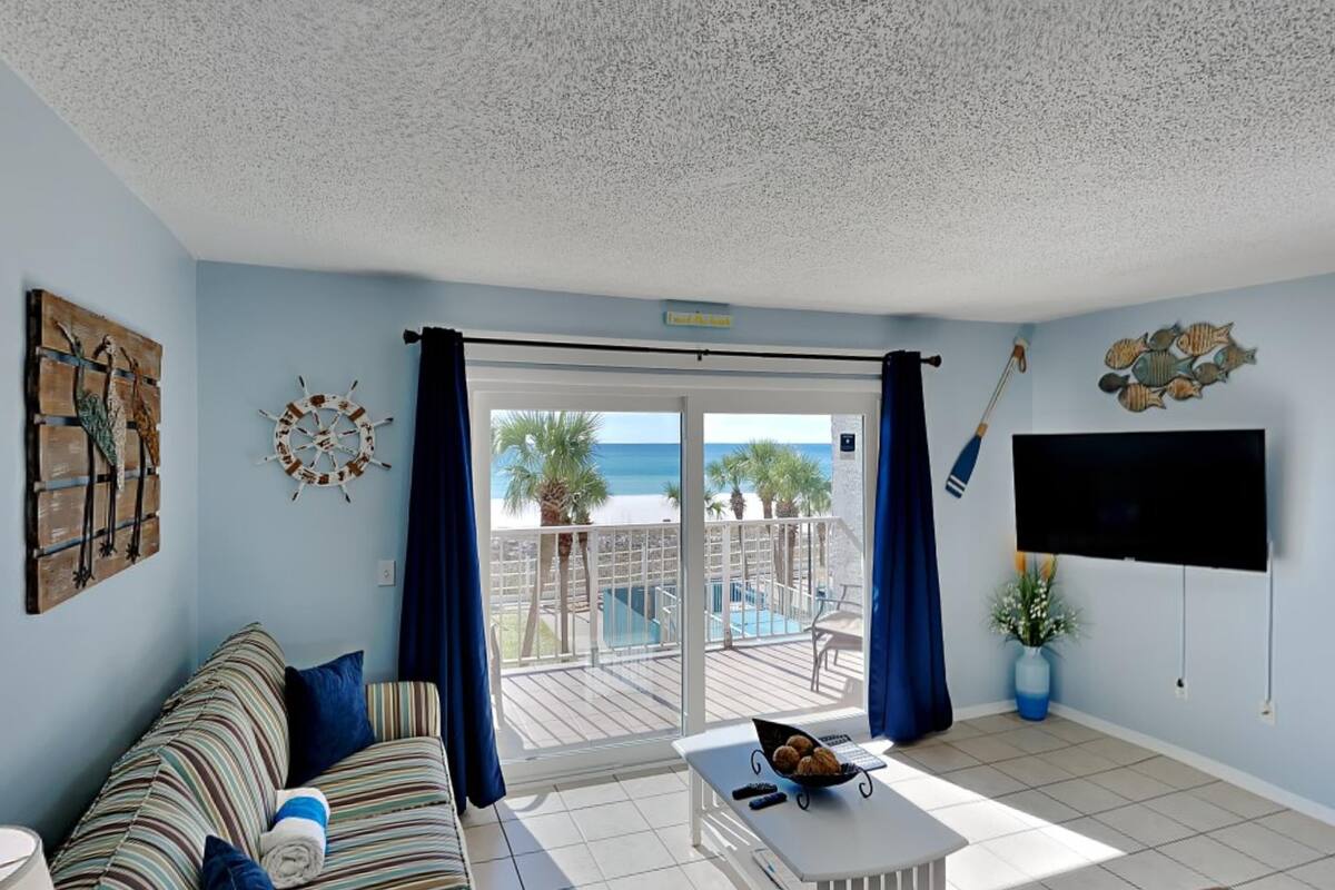 A living room with a sofa, a coffee table, and a TV. There is a balcony with a view of the beach.