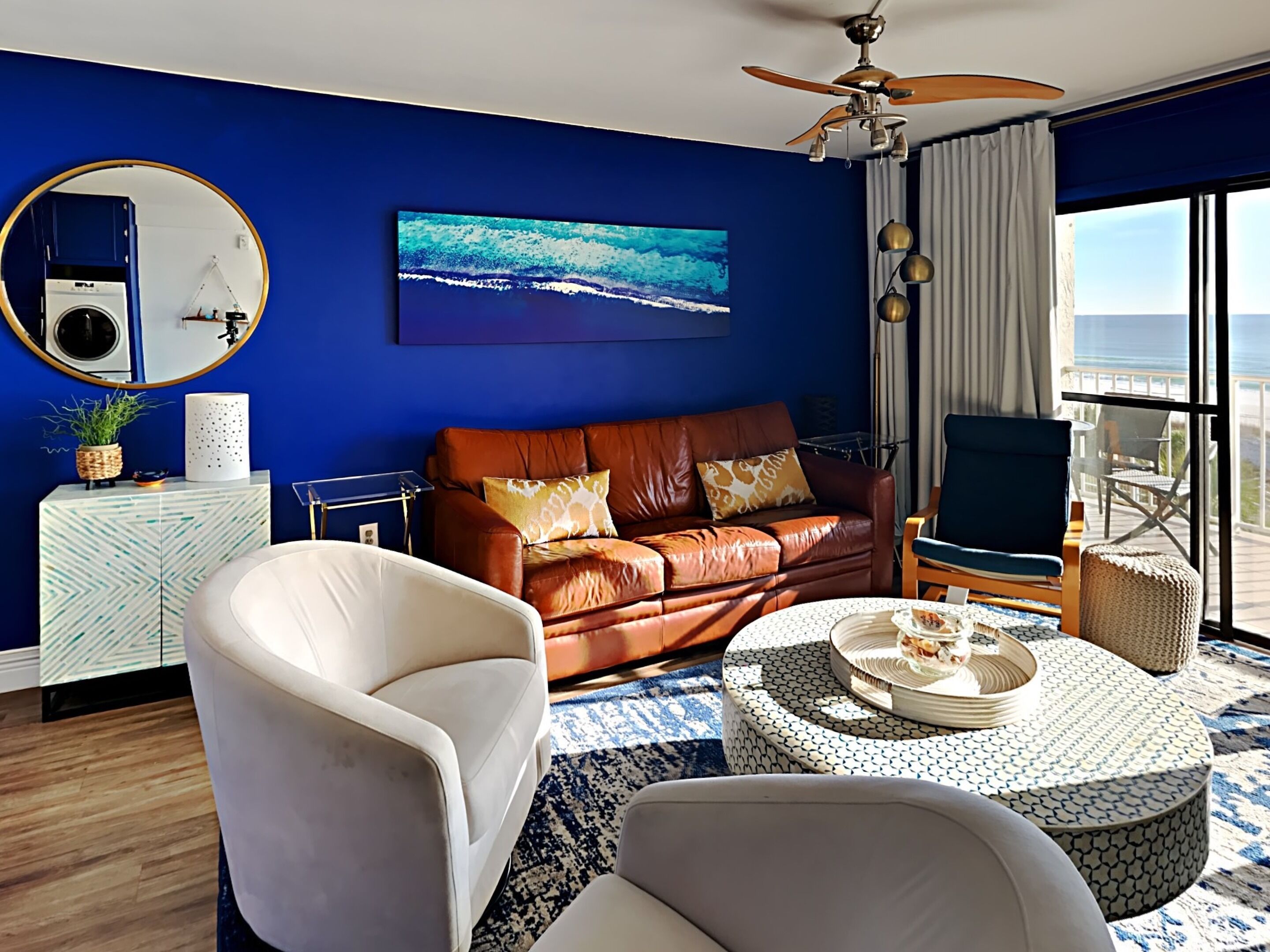 A room with a blue wall, a brown leather sofa, a white armchair, a round coffee table, and a view of the ocean through large windows.