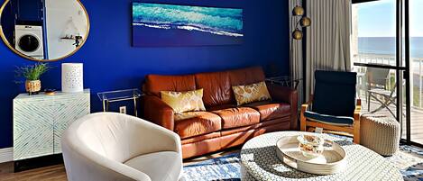 A room with a blue wall, a brown leather sofa, a white armchair, a round coffee table, and a view of the ocean through large windows.