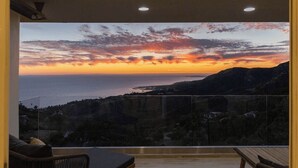 Ein Zimmer mit großem Fenster mit Blick auf eine Küstenlandschaft bei Sonnenuntergang.