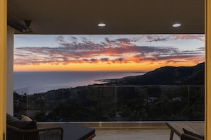 Quarto com uma janela ampla que dá para a paisagem costeira ao pôr do sol.