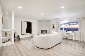 Uma sala de estar moderna com um sofá branco, lareira, escrivaninha com cadeira e uma janela grande com cortinas.