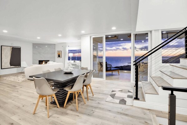 A modern living room with a dining area, large windows with a view of the ocean, and a staircase.