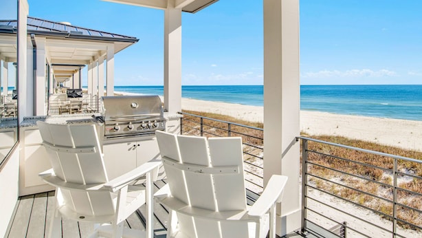 A covered outdoor area with a grill and white chairs, overlooking a beach.