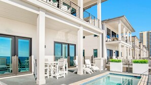 A rooftop pool area with lounge chairs and tables, a clear view of the ocean, and a modern building structure.