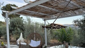 An outdoor patio with a glass table, white cushioned bench, wicker chair, and a pergola with a thatched roof. - Finca Las Fuentezuelas (Carataunas)
