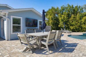 Un patio con mesa y sillas, una piscina y una casa con un ventanal grande y un cuadro.