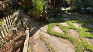 Una casa con un sendero de piedra que lleva a una terraza de madera, rodeada de abundante vegetación y un jardín cercado.