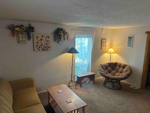 A living room with a sofa, a wooden table, a chair, and a lamp. There are wall decorations and a window with curtains.