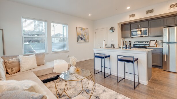 A modern living room with a sofa, a glass coffee table, and a kitchen area with a bar and stainless steel appliances. - 1 BR 1 BA Luxury - Museum District & Downtown HTX (Houston)