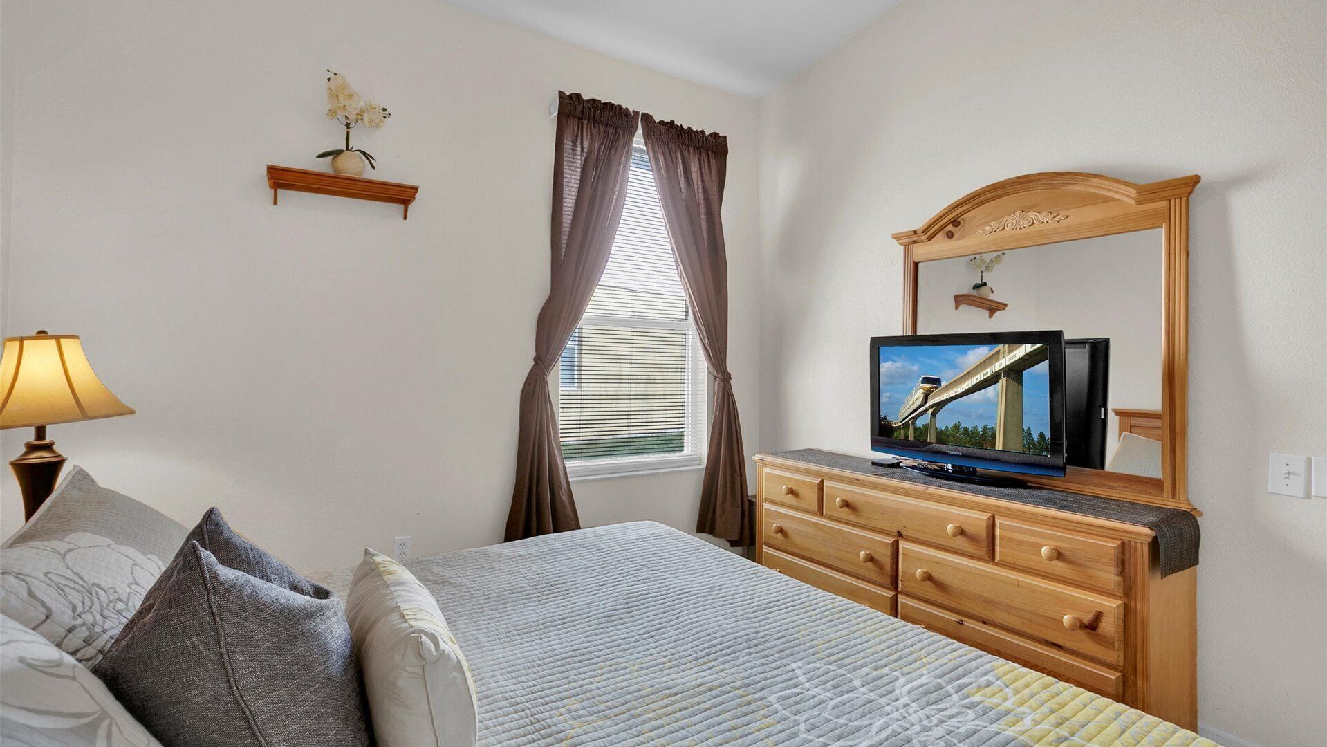 A bedroom with a bed, a dresser, a television, a window with curtains, and a wall-mounted shelf with a flower.