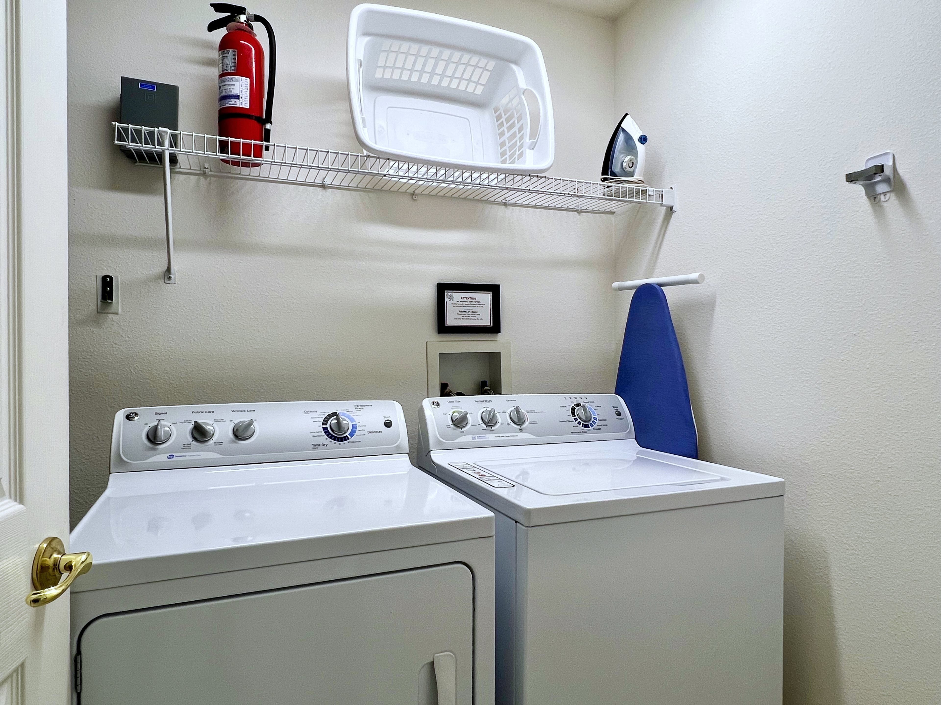 A laundry room with two washing machines, a fire extinguisher, a shelf with a laundry basket, and a wall-mounted ironing board.