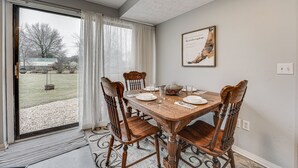 Ein Esszimmer mit Holztisch, gedeckt für vier Personen, einem großen Fenster mit Vorhängen und einem gerahmten Bild an der Wand.