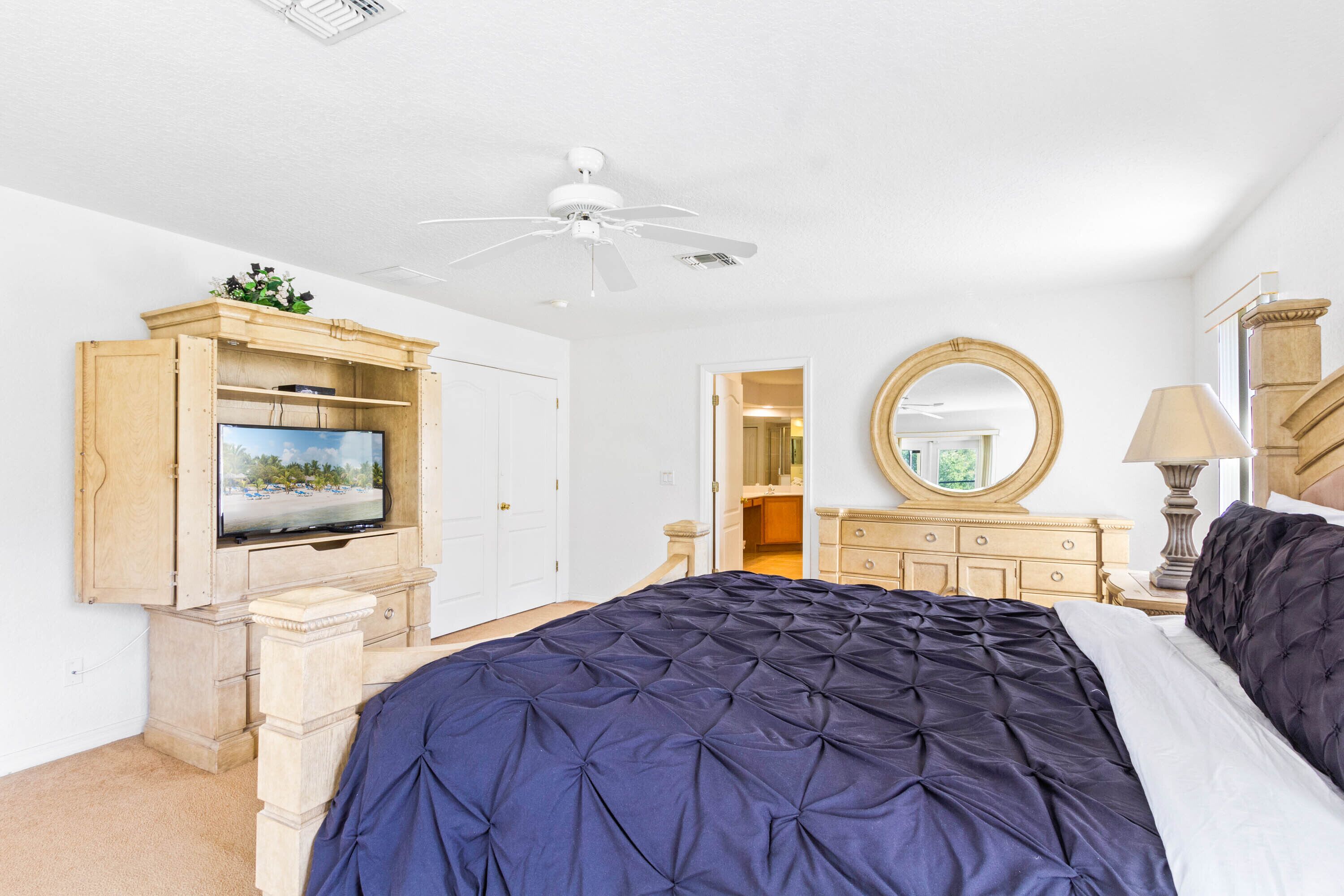 A bedroom with a large bed, a ceiling fan, a wooden dresser with a television, and a mirror.