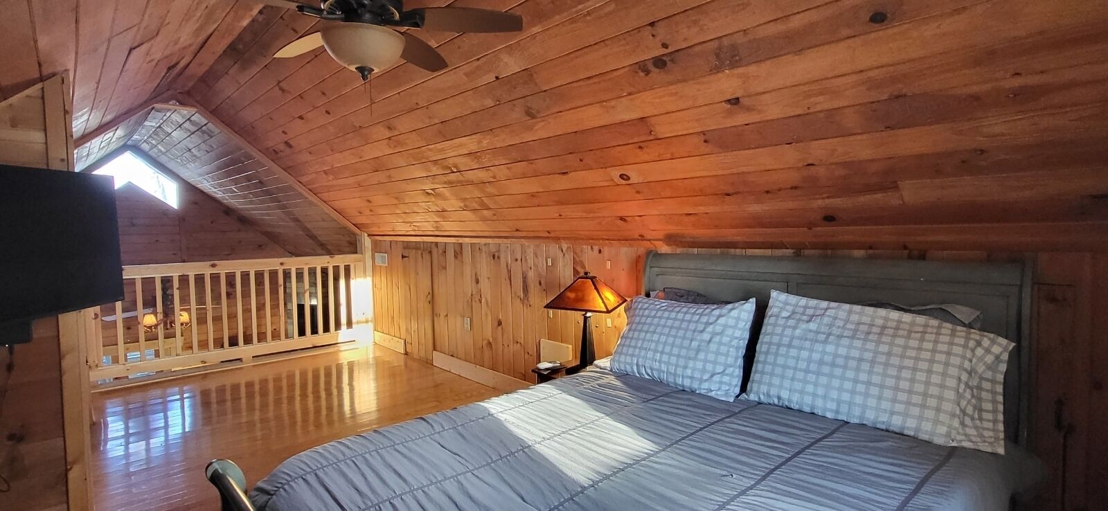 A cozy attic bedroom with a wooden ceiling, a bed with checkered bedding, a nightstand with a lamp, and a view of the balcony.