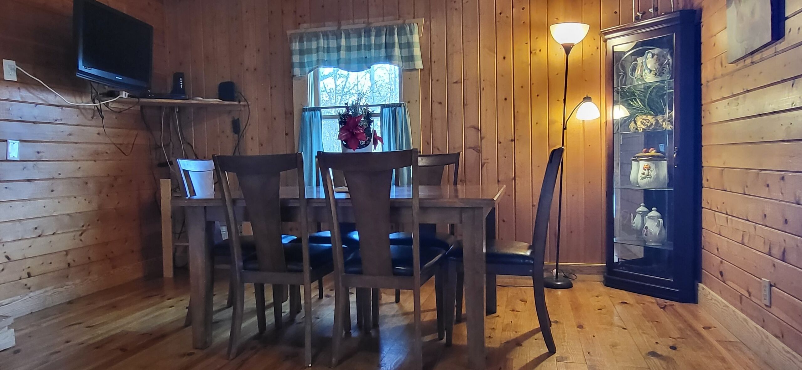 Ein Zimmer mit Holzwänden, einem Esstisch und Stühlen, einem an der Wand montierten Fernseher, einem Fenster mit Vorhängen und einer Lampe.