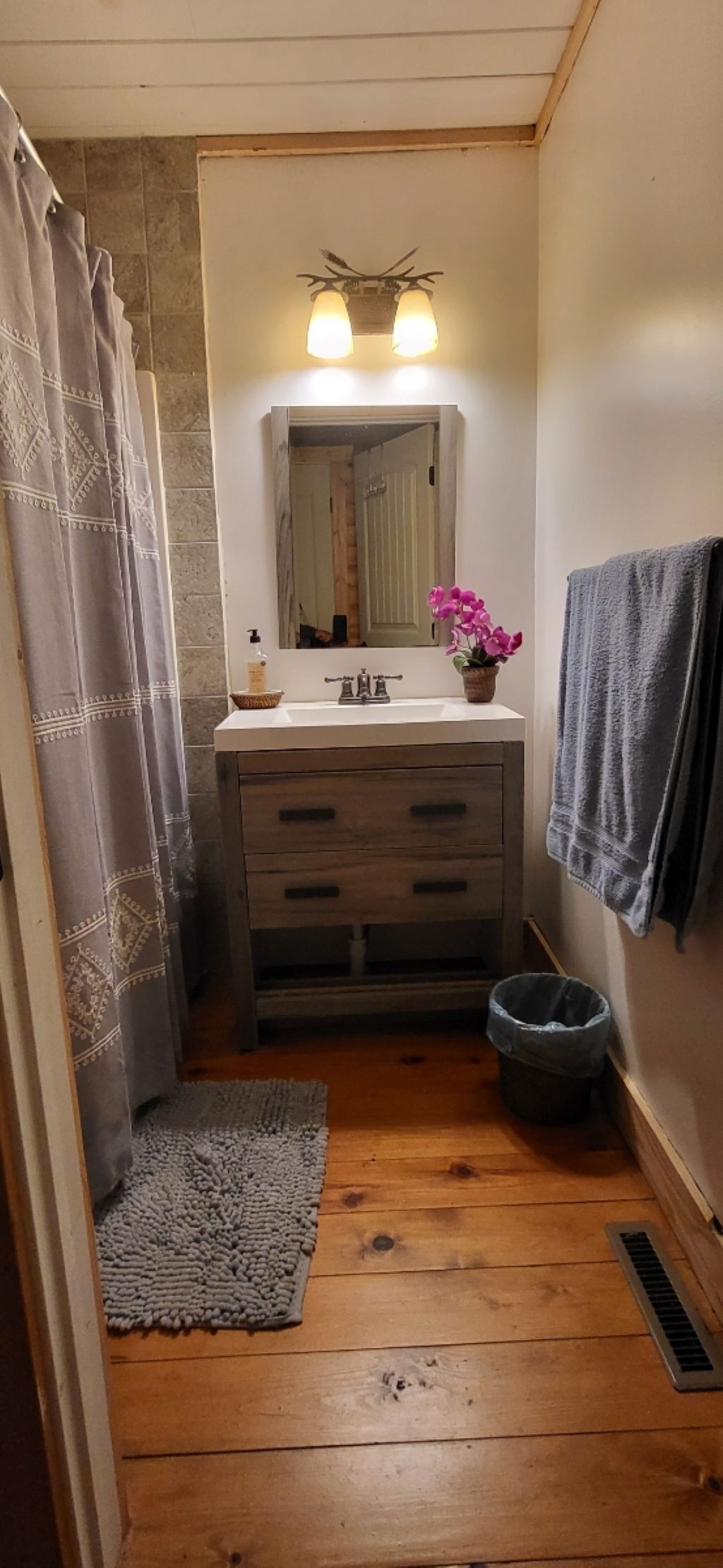 A narrow bathroom with a wooden floor, a white sink with a mirror above it, a towel hanging on the wall, and a trash bin.