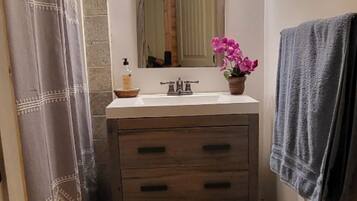 A narrow bathroom with a wooden floor, a white sink with a mirror above it, a towel hanging on the wall, and a trash bin.