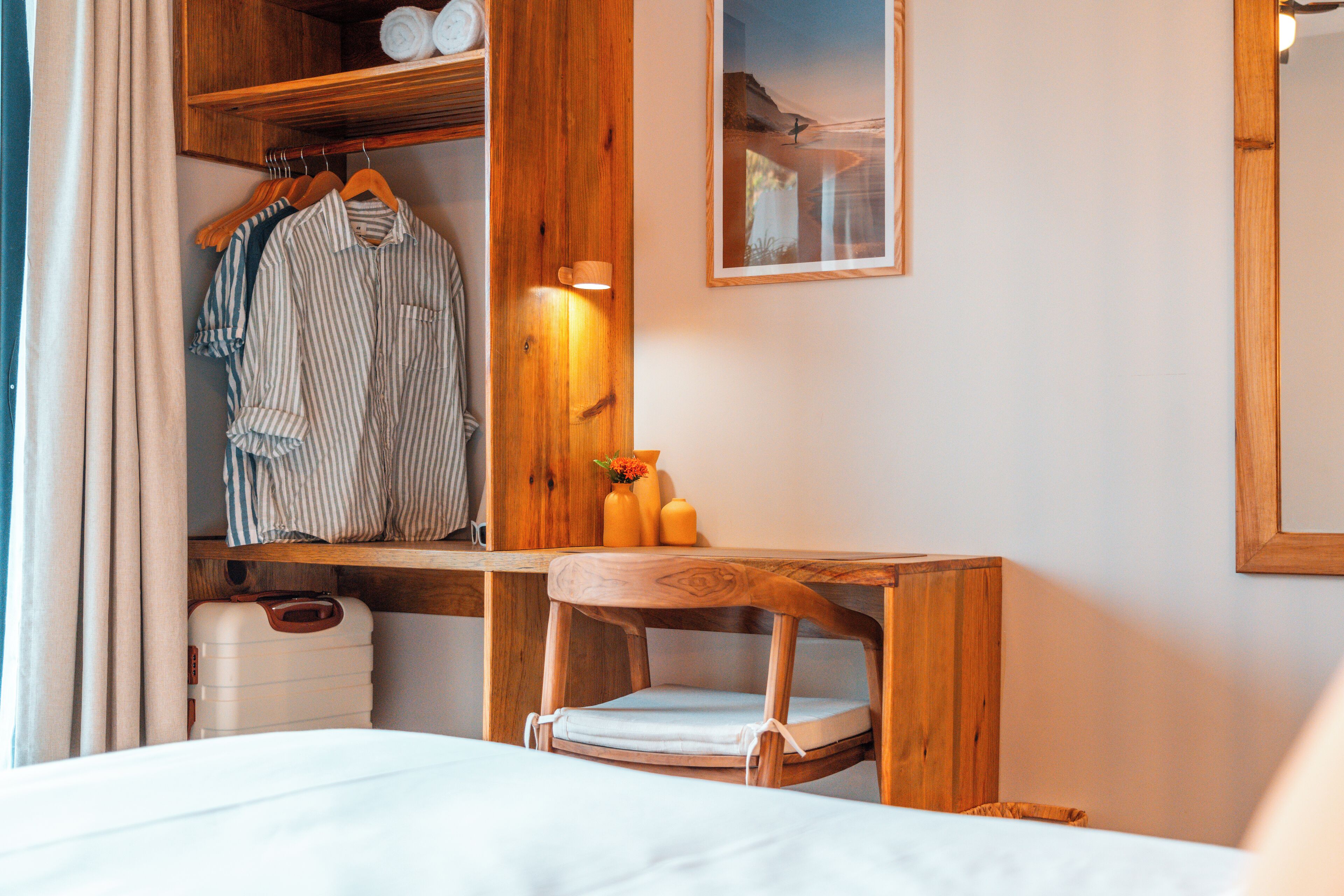 A hotel room with a wooden desk, a chair, a bed, a wardrobe, and a framed picture on the wall.