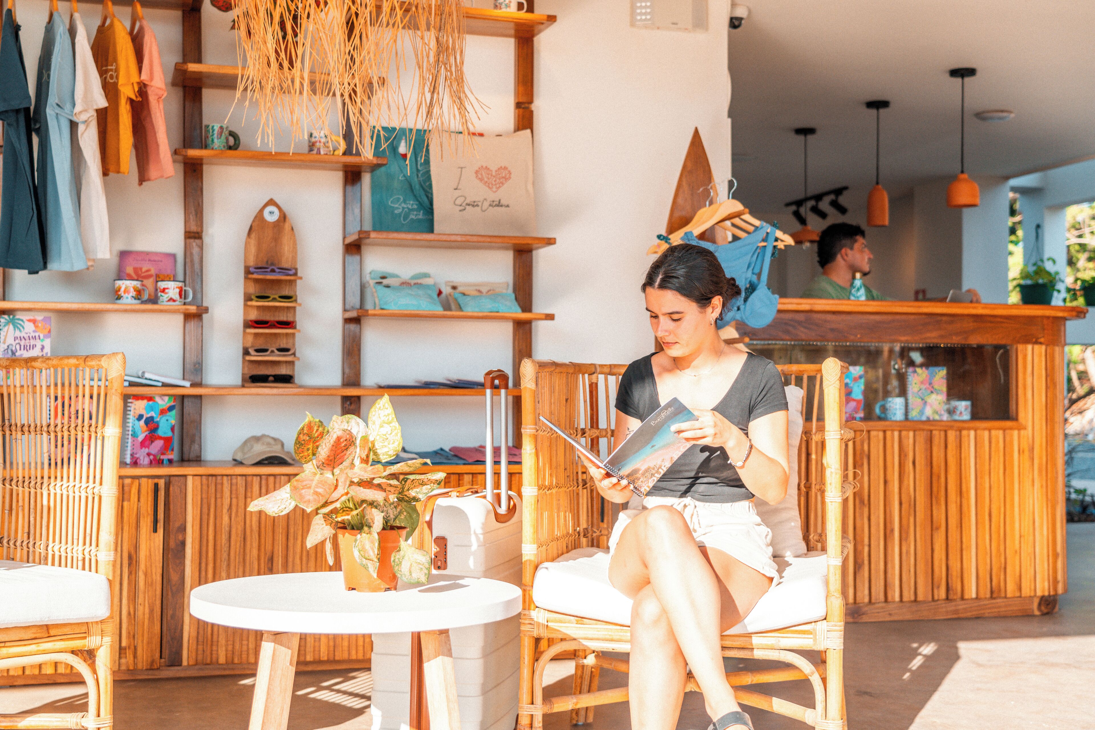 A woman sitting at a table reading a book in a cafe with wooden furniture and shelves with various items.