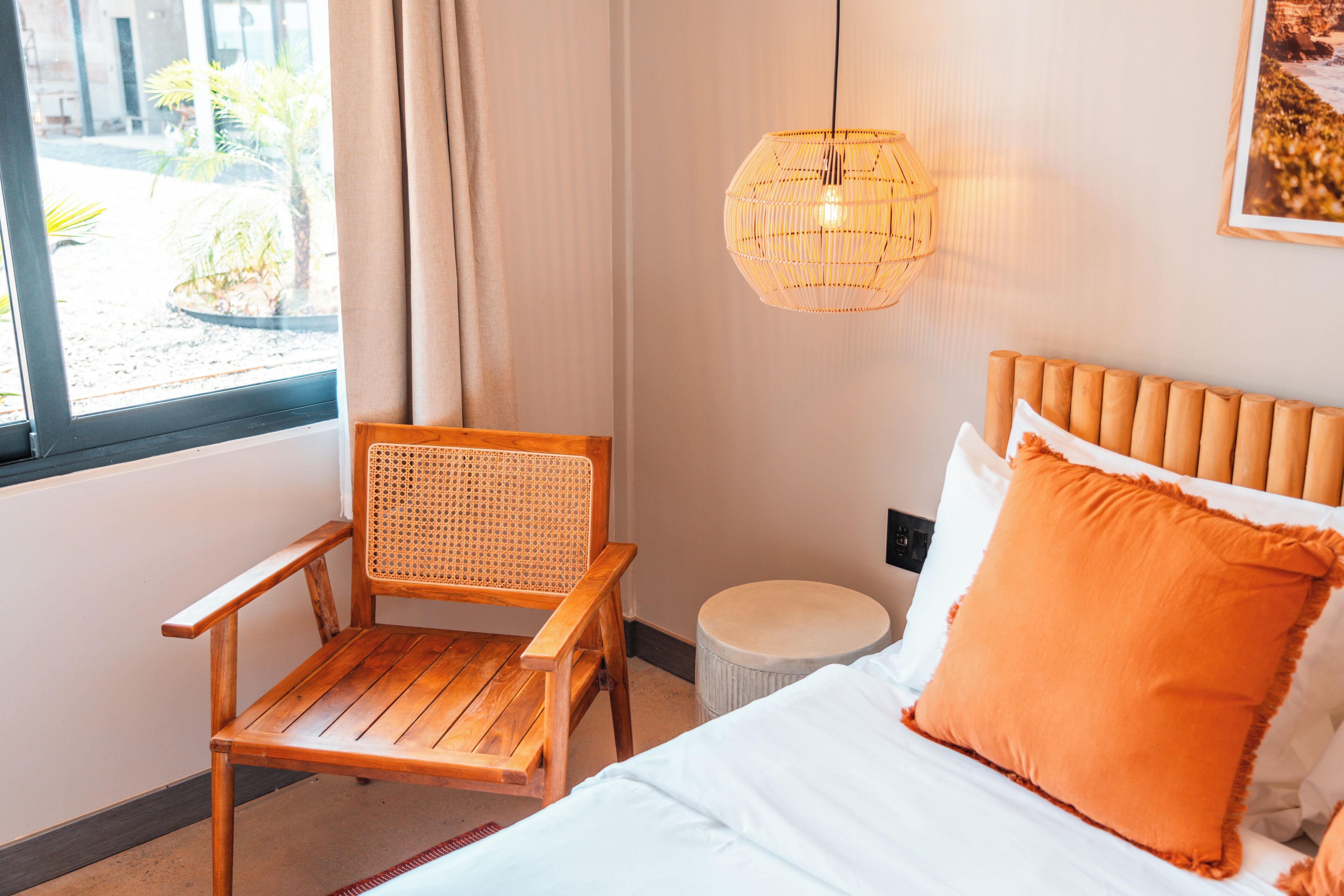 Una habitación con una cama, una silla de madera, un taburete redondo, una lámpara colgante y una ventana con cortinas.