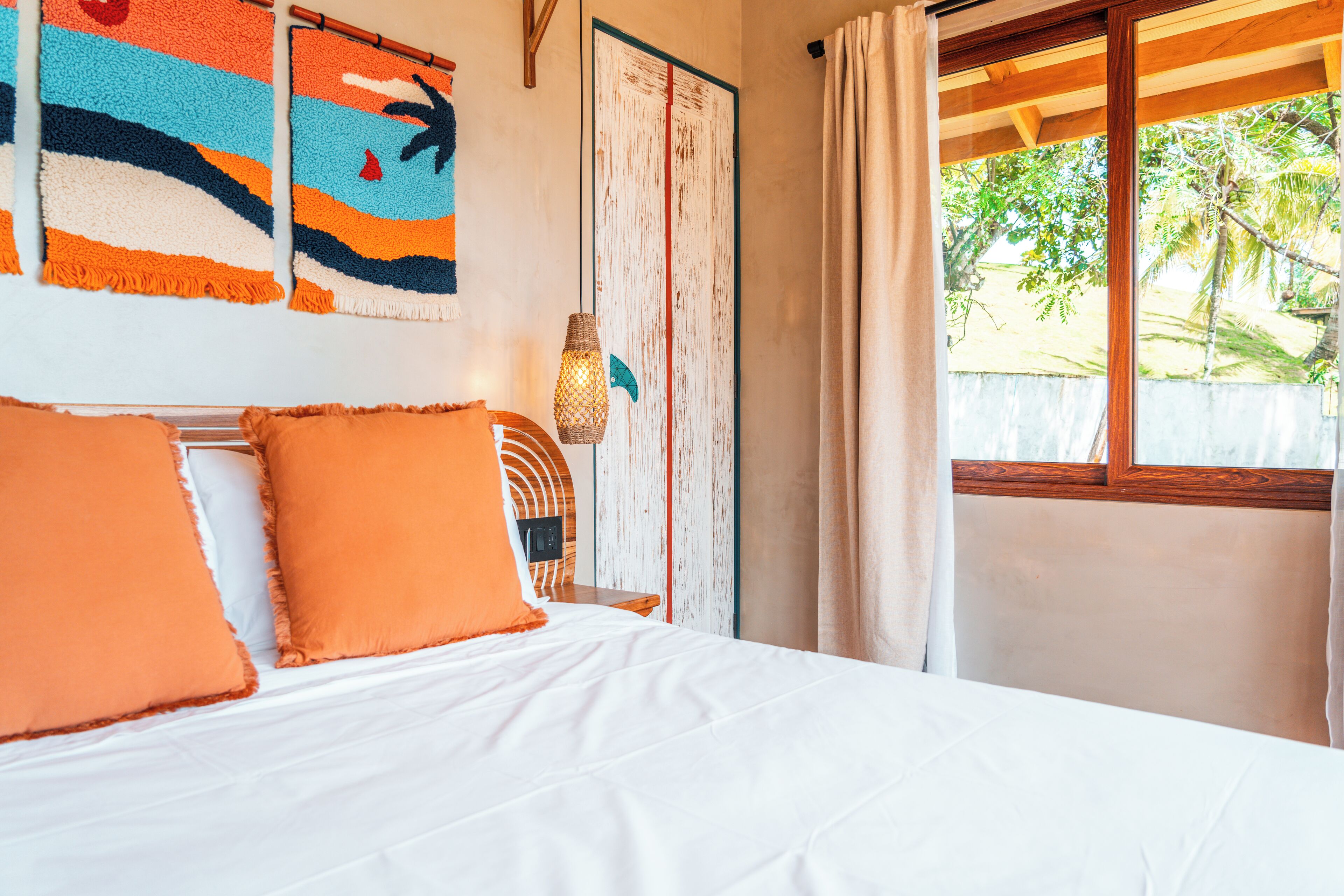 A bed with white bedding and orange pillows, a wooden headboard, a wall-mounted lamp, and a view of palm trees through a window.