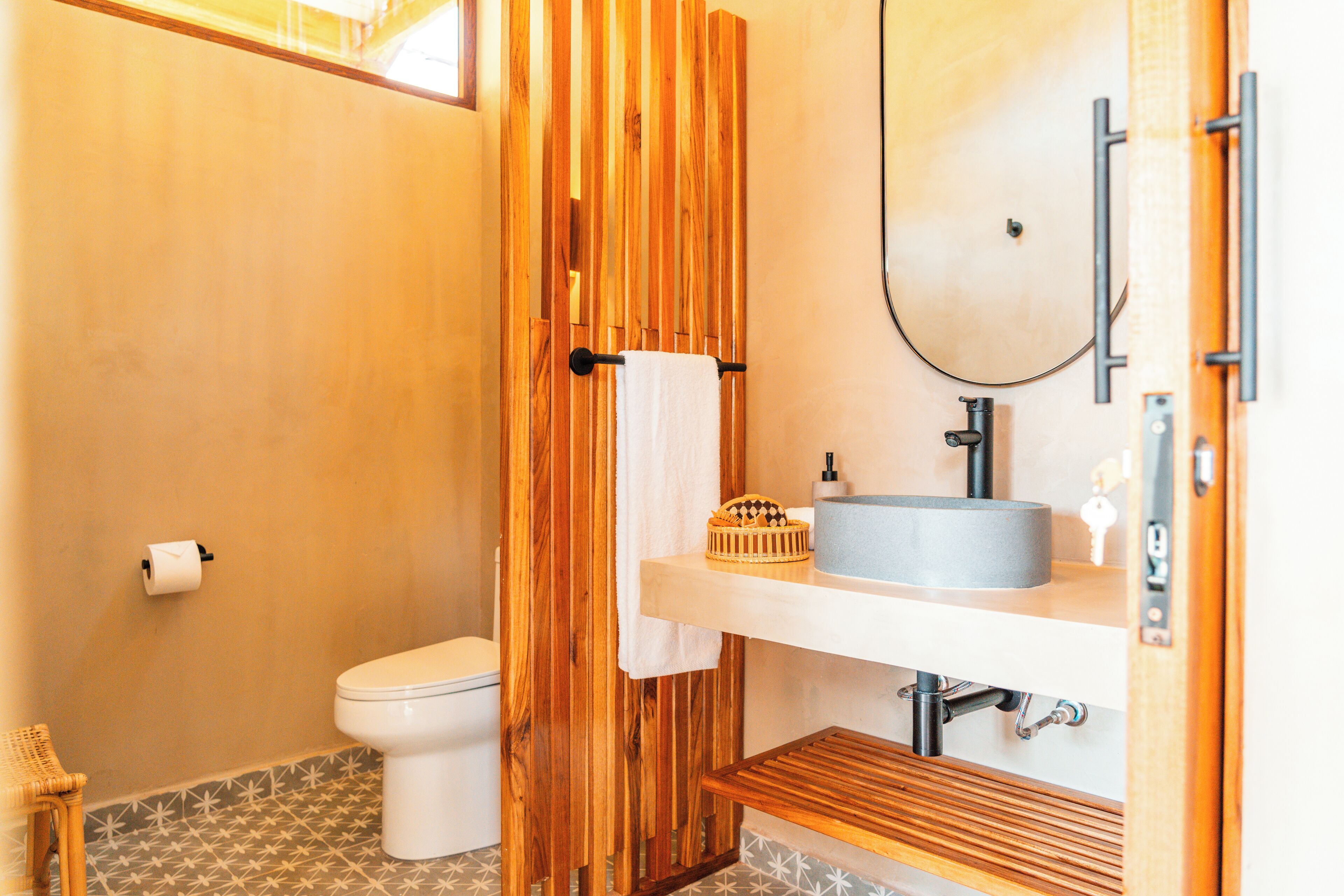 A bathroom with a wooden partition, a toilet, a sink with a mirror, and a small shelf with toiletries.