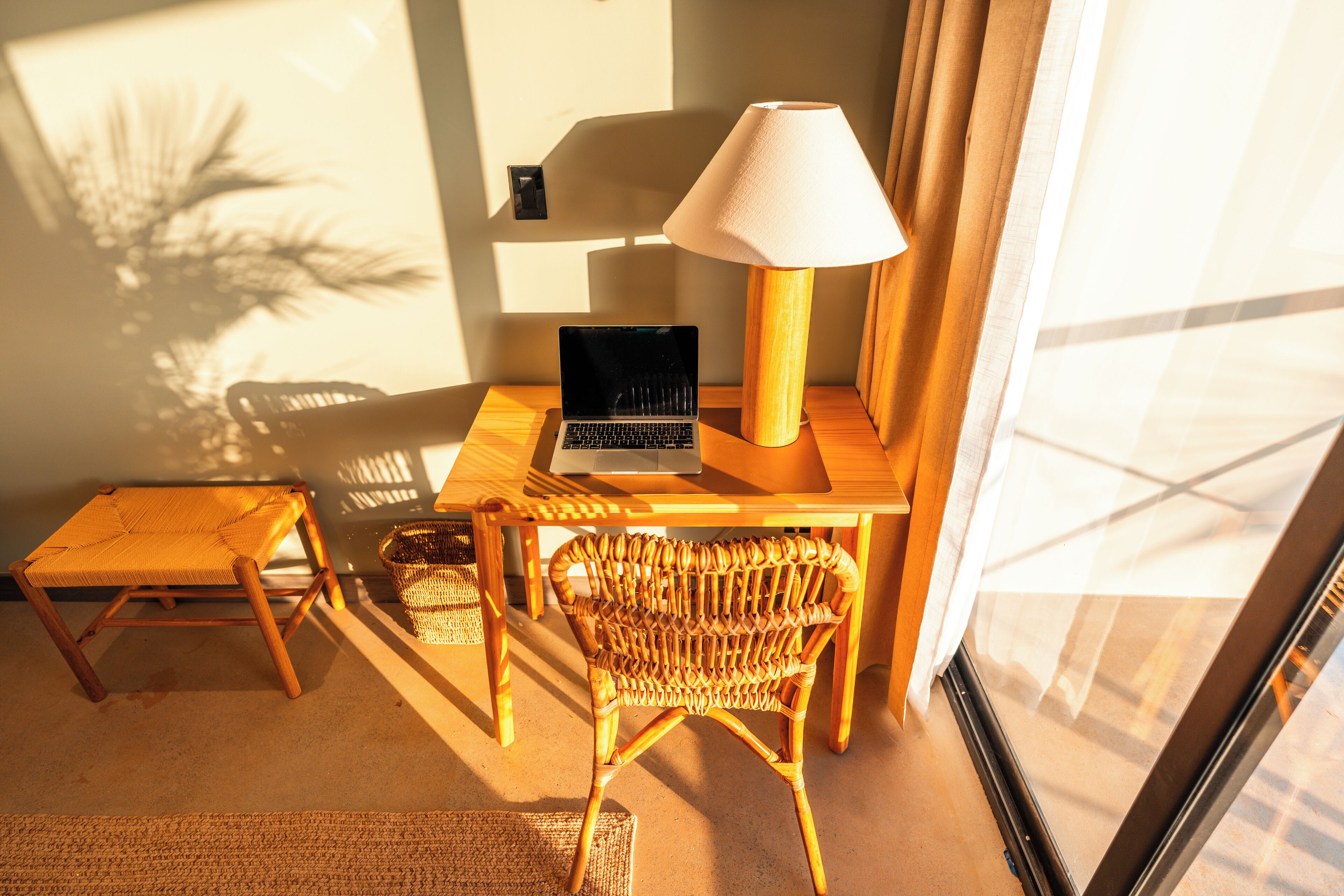 A room with a laptop on a wooden table, a wicker chair, a small stool, and a lamp.