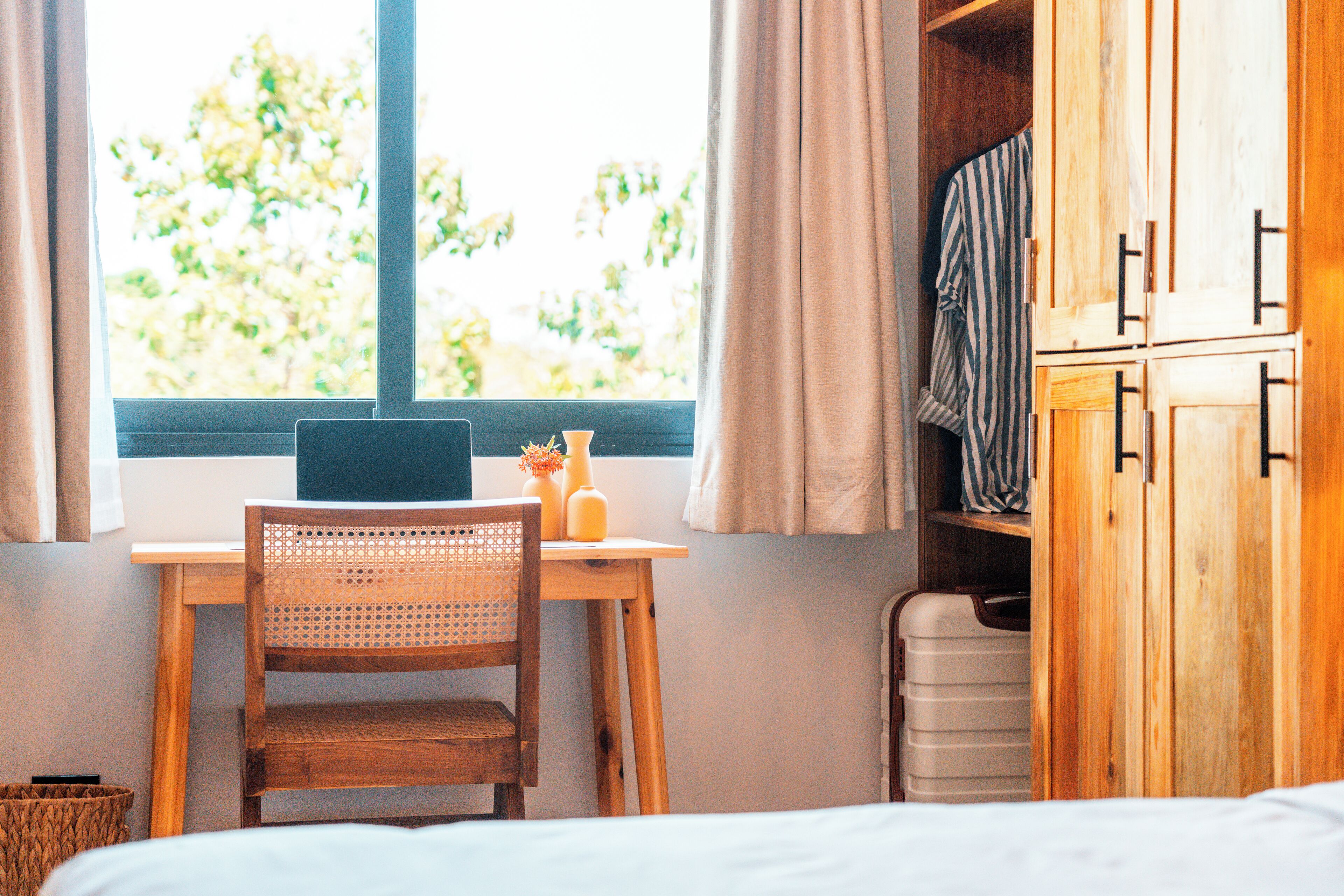 A room with a wooden desk, a laptop, a wicker chair, a suitcase, and a wardrobe.