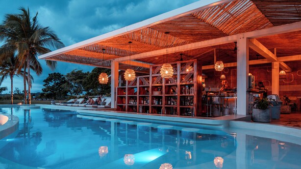 Un área moderna junto a la piscina con bar y asientos tipo lounge, rodeada de palmeras.