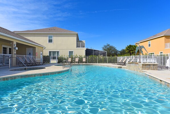 A swimming pool with lounge chairs and a covered area, surrounded by buildings with a fence.