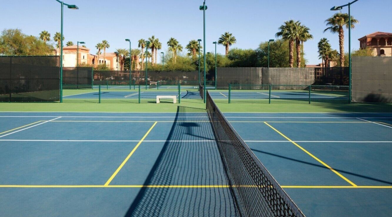 Ein Tennisplatz im Freien mit mehreren Spielfeldern, Bänken und Palmen im Hintergrund.